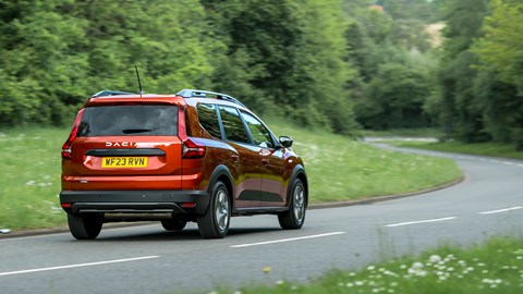 Dacia Jogger Hybrid (2023): rear three quarter driving, wide angle, country lane, orange car