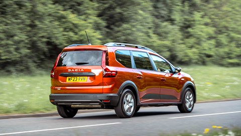Dacia Jogger Hybrid (2023): rear three quarter driving, fly-by, wood in background, orange car