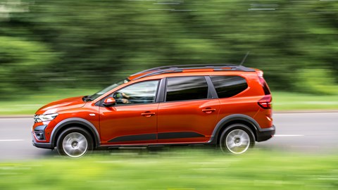 Dacia Jogger Hybrid (2023): side view quarter driving, fly-by, wood in background, orange car