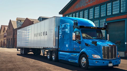 A Waymo branded HGV with a blue cab and white trailer sits parked in front of a warehouse