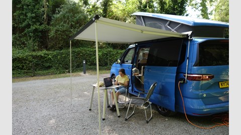 Image of a woman reading while sat under the awning of a blue Volkswagen California