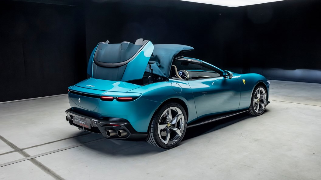 Ferrari Ferrari: Vista trasera de un Ferrari Amalfi Spider en color celeste, capturado durante la operación de plegado de su techo rígido retráctil (RHT) de lona negra, mostrando la tapa del compartimento del techo abierta y el mecanismo de cinemática compleja en acción para guardar el panel sobre el eje posterior, priorizando la aerodinámica y la distribución de pesos.